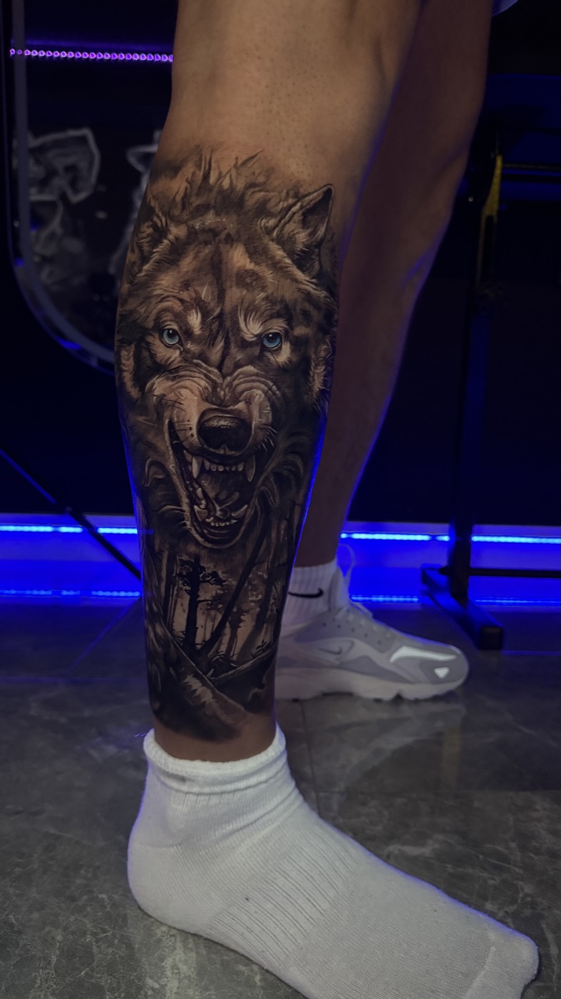 Wolf tattoo on the lower leg with bared teeth, detailed fur and a forest beneath the face